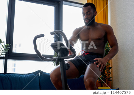 fit muscular man with naked torso riding a stationary bike at home, training workout 114407579