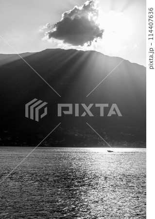 A tranquil scene capturing the serene beauty of a mountain lake at sunset. Sun rays pierce through a cloud, casting illuminating beams and sparkling reflections on the water surface. A lone boat 114407636