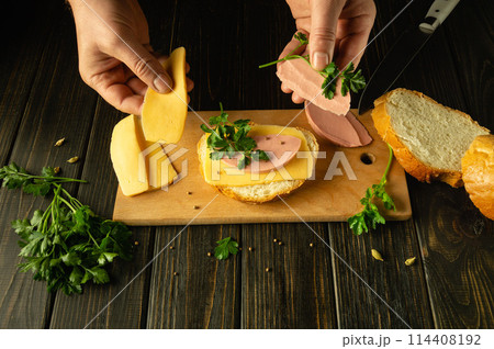 A man is preparing a delicious sandwich on a wooden board. Low key concept of preparing a sandwich with sausage and cheese on the kitchen table A man is preparing a delicious sandwich on a wooden board. Low key concept of preparing a sandwich with sausage and cheese on the kitchen table 114408192