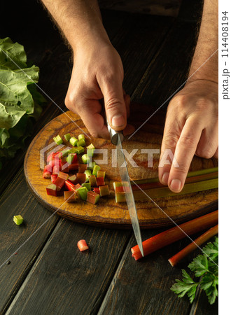 Cutting rhubarb with a knife in the hand of a chef for preparing a vegetarian dish on the kitchen table. Place for advertising on a dark background 114408194
