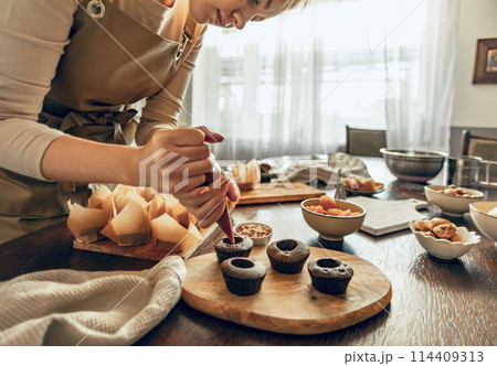 Woman skillfully decorates cupcakes on a wooden board, surrounded by baking supplies and kitchen tools, showcasing the artistry of homemade desserts Woman skillfully decorates cupcakes on a wooden board, surrounded by baking supplies and kitchen tools, showcasing the artistry of homemade desserts 114409313
