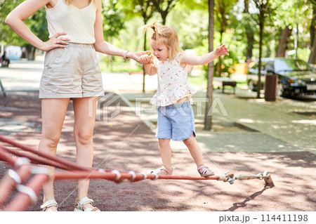 Active little child playing on climbing net and jumping on trampoline at school yard playground. Kids play and climb outdoors on sunny summer day. Cute girl on nest swing at preschool sport center. 114411198