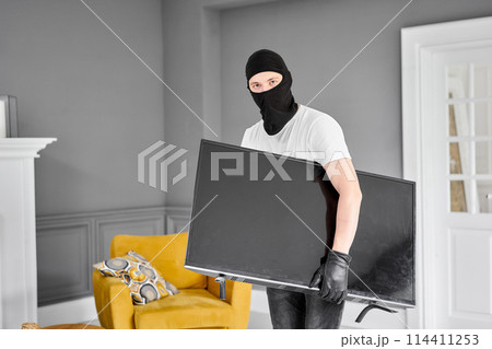 Thief with black balaclava stealing modern expensive television. Masked face. Man burglar stealing tv set from house Thief with black balaclava stealing modern expensive television. Masked face. Man burglar stealing tv set from house 114411253