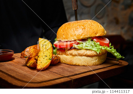 Burger with chicken cutlet, wheat bun, Cheddar cheese, tomatoes, lettuce and creamy mustard sauce, with a knife stuck into it, with baked potatoes 114411317