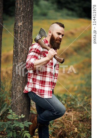 bearded lumberjack worker standing in forest with axe 114411978