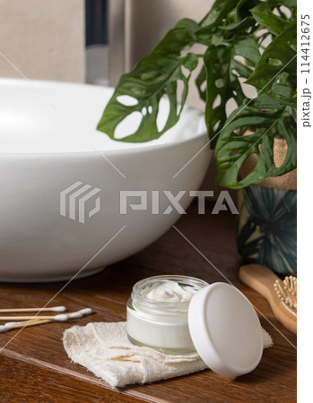 Opened cosmetic cream jar near basin and green plant on wooden countertop in bath, lid mockup 114412675