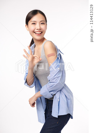 young asian women smiling after getting a vaccine, holding down her shirt sleeve and showing her arm with bandage after receiving vaccination on white background, and she shows ok sign, 114413129