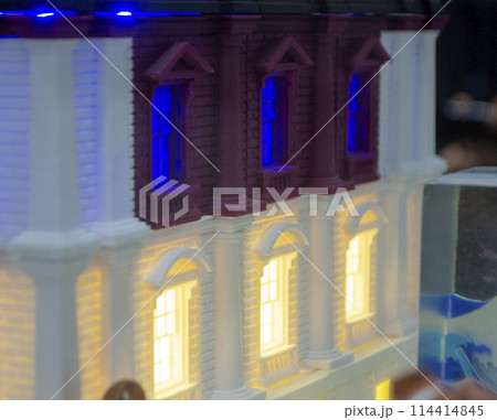 Prototype printed on 3D printer brick building with columns and glowing windows. Model of building with white and brown color, windows with light shining, created by 3D printer from molten plastic 114414845