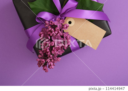 Black craft gift box with empty tag, lilac flowers on violet background. Place for your text Black craft gift box with empty tag, lilac flowers on violet background. Place for your text 114414947