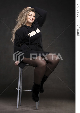 Beautiful fat woman with long curly hair in black clothes on a gray background. Plus size model girl. 114415097