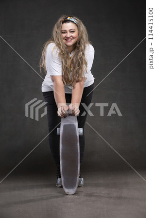 Beautiful fat woman with a skateboard on a gray background looks at the camera and smiles. Beautiful fat woman with a skateboard on a gray background looks at the camera and smiles. 114415100