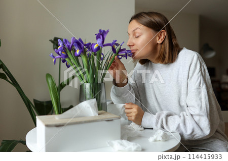 Seasonal allergy free. Young woman sniffs of iris flowers, enjoys of smell without of runny nose, itching or cough seasonal symptoms at cozy home. Girl sits by the table. Used paper tissues on table Seasonal allergy free. Young woman sniffs of iris flowers, enjoys of smell without of runny nose, itching or cough seasonal symptoms at cozy home. Girl sits by the table. Used paper tissues on table 114415933