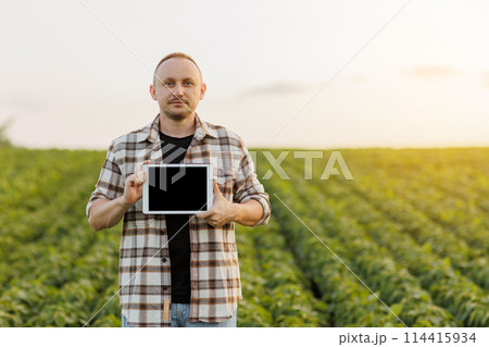 Male farmer shows digital tablet with empty black screen on green soybeans field. Smart farming technology and organic agriculture concept. Quality control growth and development soya plants Male farmer shows digital tablet with empty black screen on green soybeans field. Smart farming technology and organic agriculture concept. Quality control growth and development soya plants 114415934