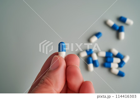 A hand holds a white and blue pill on a blue background. A hand holds a white and blue pill on a blue background. 114416303