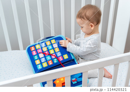 Little baby girl playing with busy book while sitting in crib. Concept of quiet books and modern educational toys 114420321