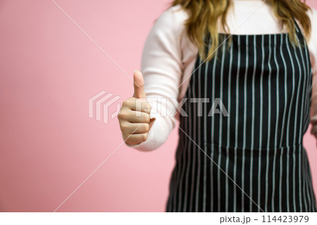 Young woman in pink sweater and striped apron give a thumbs up gesture. Portrait on pink background with studio light. 114423979