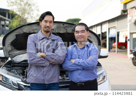 A mechanic in work uniform and senior customer, stand with their arm cross, in front of an open car hood at car dealership, satisfaction with a repair or inspecting the vehicle service. 114423981