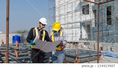 Two construction worker looking at a floor plan, standing in front of a large building site with helmet and safety gear. Day time work safety check. Work environment at the site of housing projects. Two construction worker looking at a floor plan, standing in front of a large building site with helmet and safety gear. Day time work safety check. Work environment at the site of housing projects. 114423987