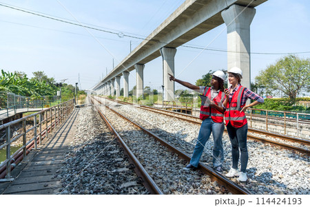 Two construction or engineer with safety helmet and high-visibility vest reviewing work to be done or progress on a railway track project while holding tablet computer and walkie-talkie. 114424193