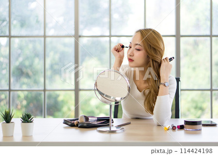 A woman is applying makeup while looking into a round mirror on a table. She is focused on applying mascara and has a collection of makeup items and brushes in front of her as well as a makeup palette 114424198