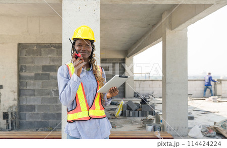 A builder in safety uniform standing at a building site, holding a walkie-talkie communicating with other team member. Tablet in her hand used for reviewing construction plan or checking schedule. 114424224
