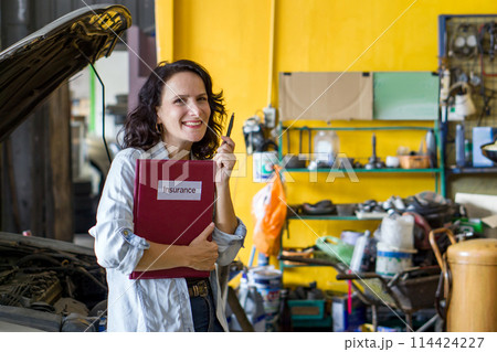 A woman standing in an auto repair shop, smiling and holding a red folder with the word "Insurance" written on the cover. There is a pen in her hand. A car with its hood open is in the background. 114424227