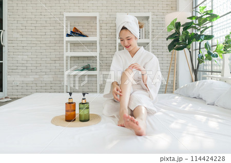 Young asian woman in a white bathrobe and towel on head, sit on a bed with lotion bottle nearby. Spreading body lotion cream gel on leg and knee after showering and taking bath, healthcare 114424228