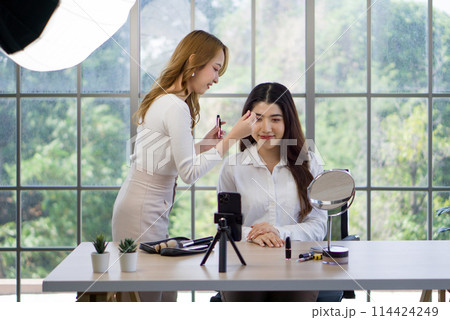 Young asian makeup artist applying makeup to the sitting woman, the subject of a beauty tutorial or makeover session, given the presence of a smartphone on a tripod presumably capturing the process. 114424249