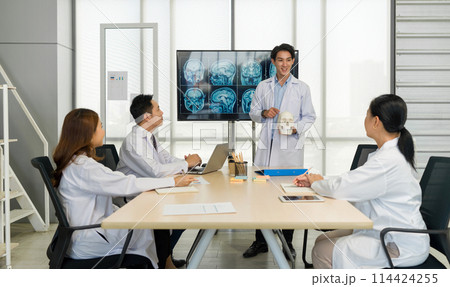 Young medical professional standing and presenting, holding a model of human skull and referencing image of brain scan displayed on a screen. The audience listening attentively with the presentation. Young medical professional standing and presenting, holding a model of human skull and referencing image of brain scan displayed on a screen. The audience listening attentively with the presentation. 114424255
