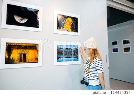 Asian woman hold camera at art gallery collection in front framed paintings pictures on wall and looking, Photographer visit at photo frame to leaning against at show exhibit artwork gallery picture 114424354