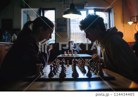 Two young individuals deeply engrossed in a strategic game of chess, competition under soft lighting Two young individuals deeply engrossed in a strategic game of chess, competition under soft lighting 114434405