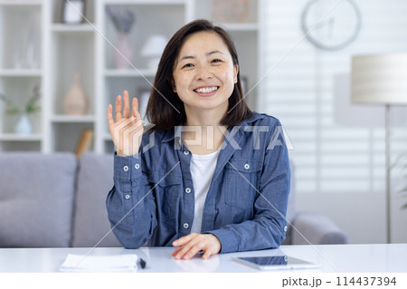 Smiling Asian woman sitting at her desk and waving at the camera. This image portrays a friendly conversation during a remote video call Perfect of communication and modern workplaces at home. 114437394