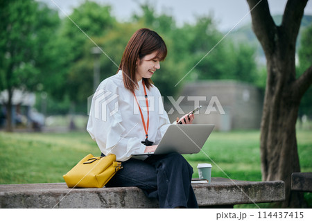 公園でパソコンとスマホを操作する若い女性 公園でパソコンとスマホを操作する若い女性 114437415