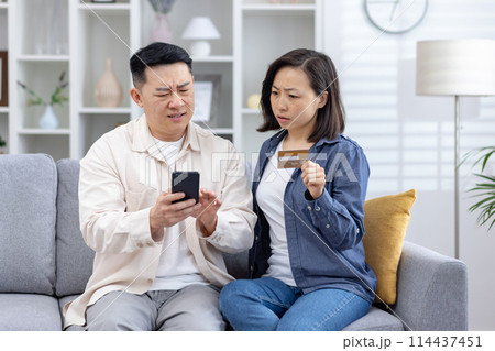 Asian couple looks distressed as they uncover unauthorized charges on their credit card phone, man confusion while the woman, holding the card, seems anxious. stress, frustration of financial fraud. 114437451