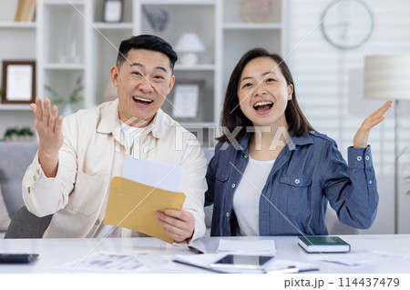 An overjoyed Asian couple in their living room, smiling and celebrating as they read some good news from a paper envelope. Their expressions reflect pure happiness and excitement. An overjoyed Asian couple in their living room, smiling and celebrating as they read some good news from a paper envelope. Their expressions reflect pure happiness and excitement. 114437479