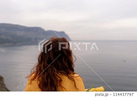 Rear view of a woman with long hair against a background of mountains and sea. Holding a bouquet of yellow tulips in her hands, wearing a yellow sweater Rear view of a woman with long hair against a background of mountains and sea. Holding a bouquet of yellow tulips in her hands, wearing a yellow sweater 114437797