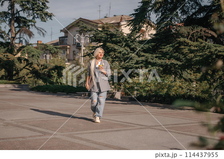 A woman is walking down a sidewalk with a cup in her hand. She is wearing a jacket and jeans. A woman is walking down a sidewalk with a cup in her hand. She is wearing a jacket and jeans. 114437955