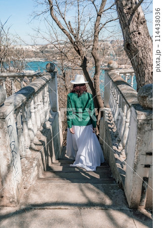 Woman walks around the city, lifestyle. Happy woman in a green jacket, white skirt and hat is sitting on a white fence with balusters overlooking the sea bay and the city. Woman walks around the city, lifestyle. Happy woman in a green jacket, white skirt and hat is sitting on a white fence with balusters overlooking the sea bay and the city. 114438036