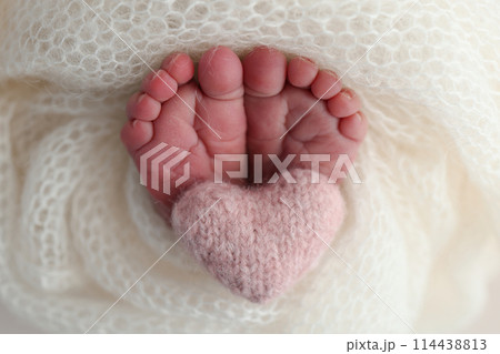 The tiny foot of a newborn baby. Soft feet of a new born in a white wool blanket. Close up of toes, heels and feet of a newborn. Knitted pink heart in the legs of a baby. Macro photography 114438813