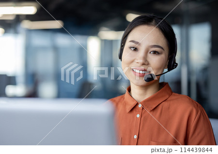 A professional woman wearing a headset, smiles warmly, displaying approachability and customer service excellence in a contemporary office environment. 114439844