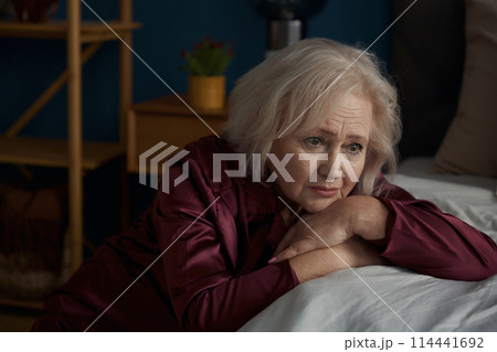 Sad elderly woman wearing pajamas sitting on bed feeling loneliness 114441692
