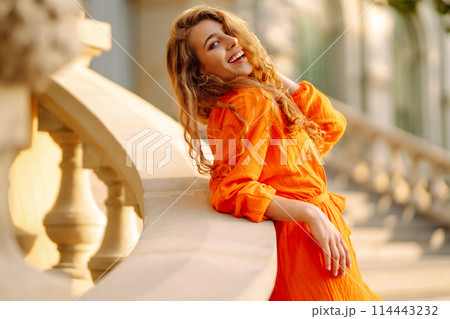 Young woman posing against backdrop of urban landscape in bright orange dress. Concept of lifestyle. 114443232