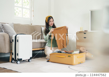 Happy Asian young woman packing suitcase at home. Preparing for summer holiday vacation abroad travel. 114447562