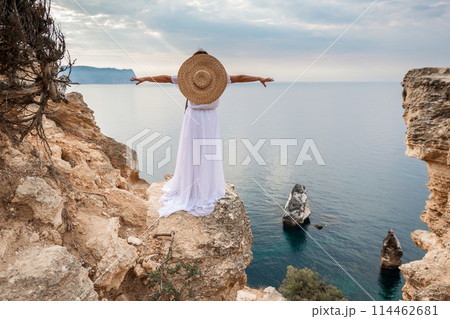 woman white dress stands on a rocky cliff overlooking the ocean. She is wearing a straw hat and she is enjoying the view. woman white dress stands on a rocky cliff overlooking the ocean. She is wearing a straw hat and she is enjoying the view. 114462681