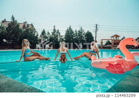 Three women are sitting in a pool, one of them is wearing a pink floatie. Scene is lighthearted and fun, as the women are enjoying their time together in the pool. Three women are sitting in a pool, one of them is wearing a pink floatie. Scene is lighthearted and fun, as the women are enjoying their time together in the pool. 114463209