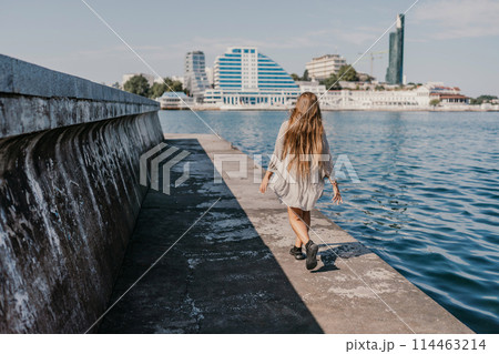 woman in a white dress is walking on a pier near the water. The scene is peaceful and serene, with the woman's long hair blowing in the wind. 114463214
