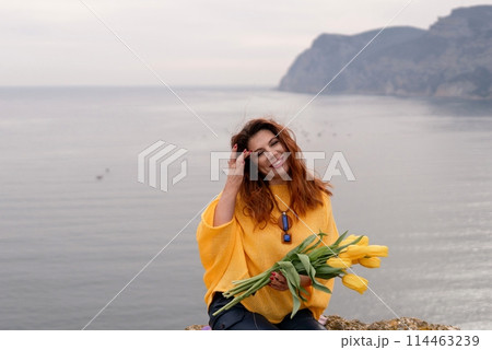 Portrait happy woman woman with long hair against a background of mountains and sea. Holding a bouquet of yellow tulips in her hands, wearing a yellow sweater 114463239