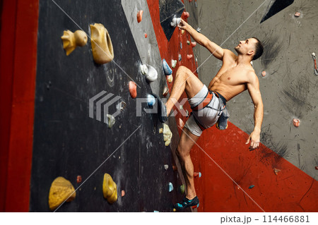 Male climber exercising in gym, climbing up on artificial wall with boulders. Male climber exercising in gym, climbing up on artificial wall with boulders. 114466881