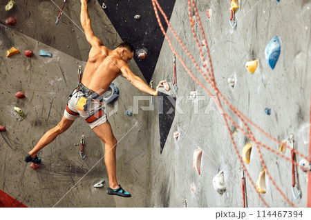 Male climber exercising in gym, climbing up on artificial wall with boulders. Male climber exercising in gym, climbing up on artificial wall with boulders. 114467394