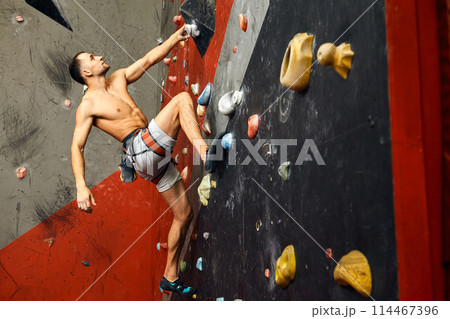 Male climber exercising in gym, climbing up on artificial wall with boulders. 114467396